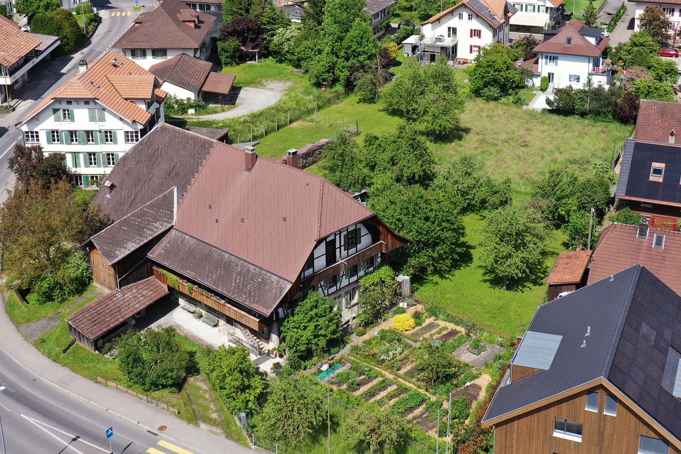 Aerial view of a residential neighborhood with a mix of traditional and modern houses, surrounded by lush greenery and gardens. The houses have red tile roofs, and some have balconies or terraces. There are also some parking spaces visible in the image.
