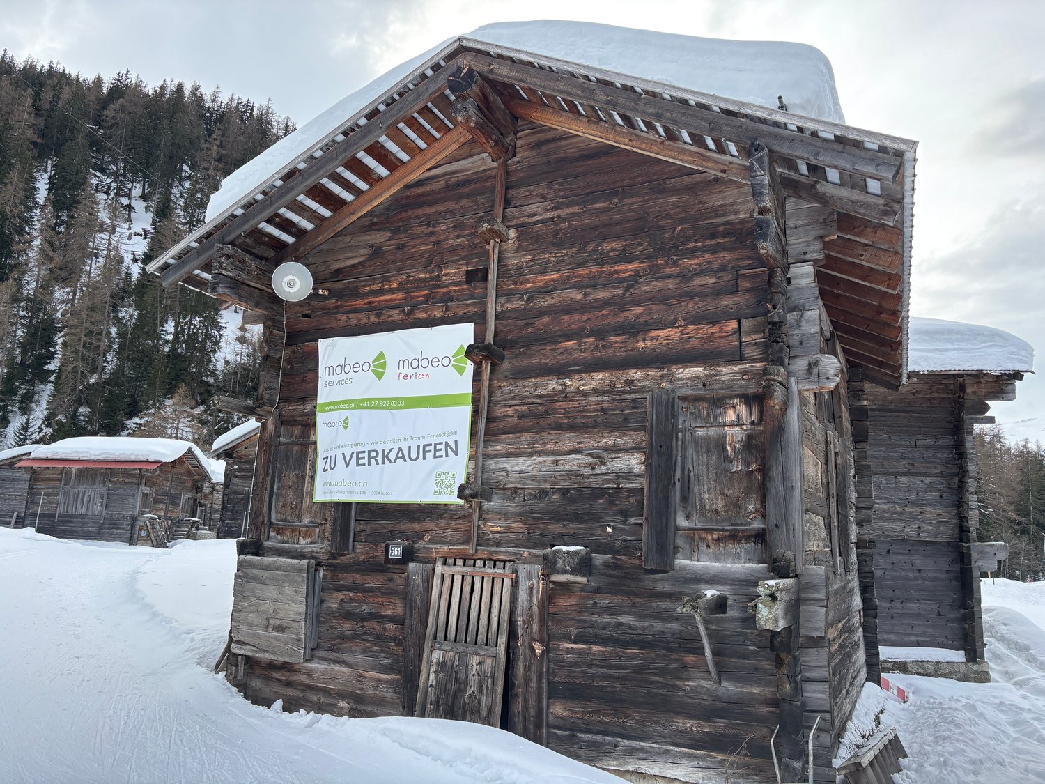 Rustic wooden cabin with a sloped roof, surrounded by snow-covered trees. The cabin has a sign indicating it is for sale.