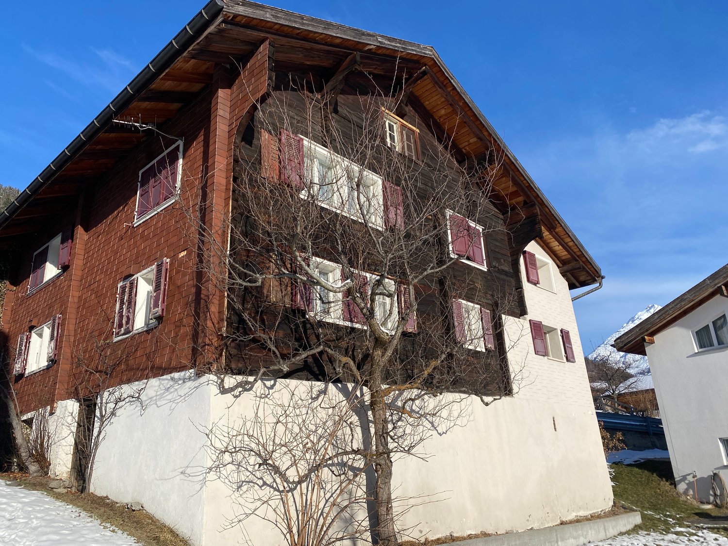 A building with a wooden wall, brick wall, and many windows. The house is surrounded by grass, and the snow is on the ground. It seems that the building is on the street. There is another building on the right, and it seems like there is a mountain in the