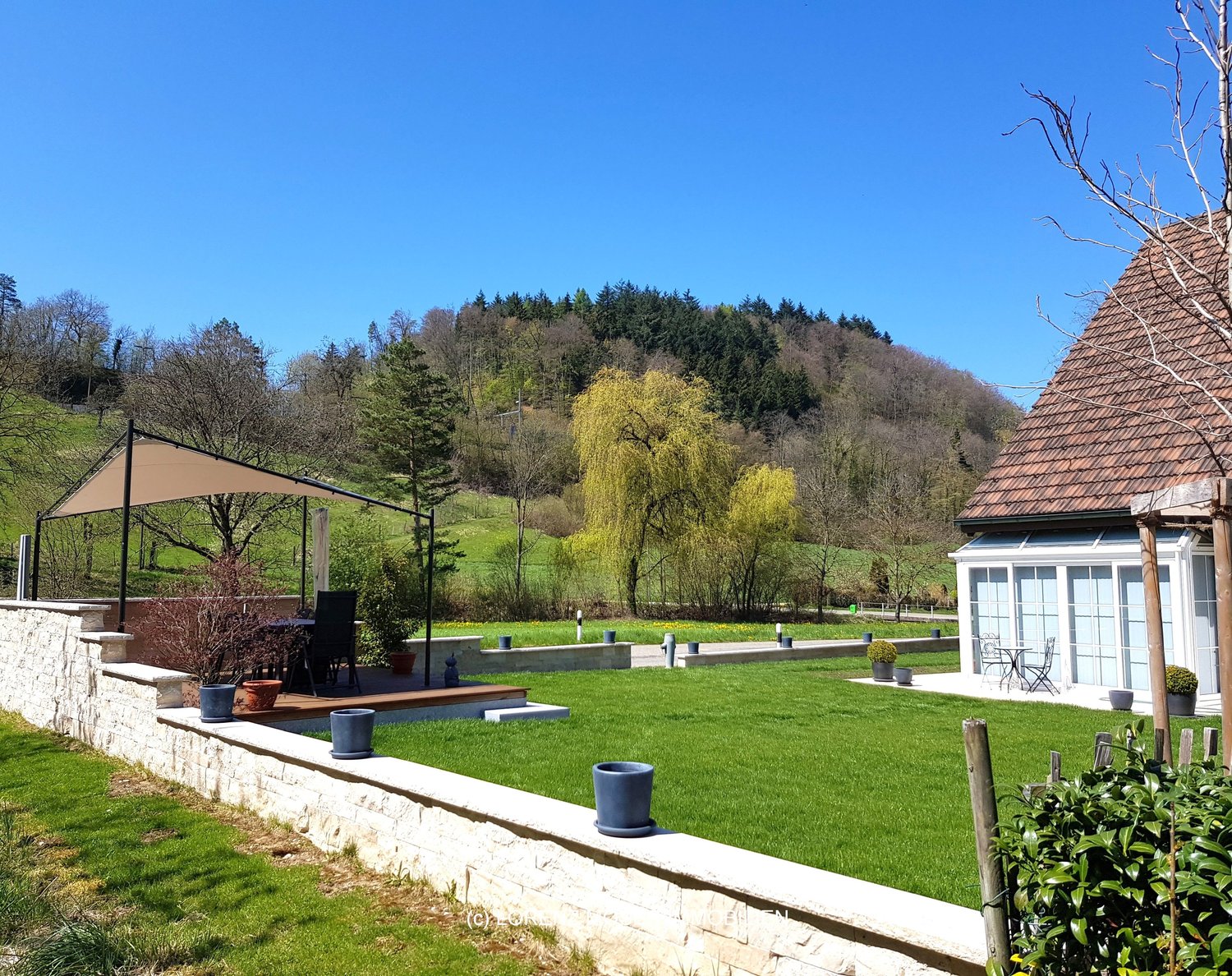 A well-maintained garden with a house in the background, stone walls, manicured lawn, terrace, potted plants, and outdoor seating.