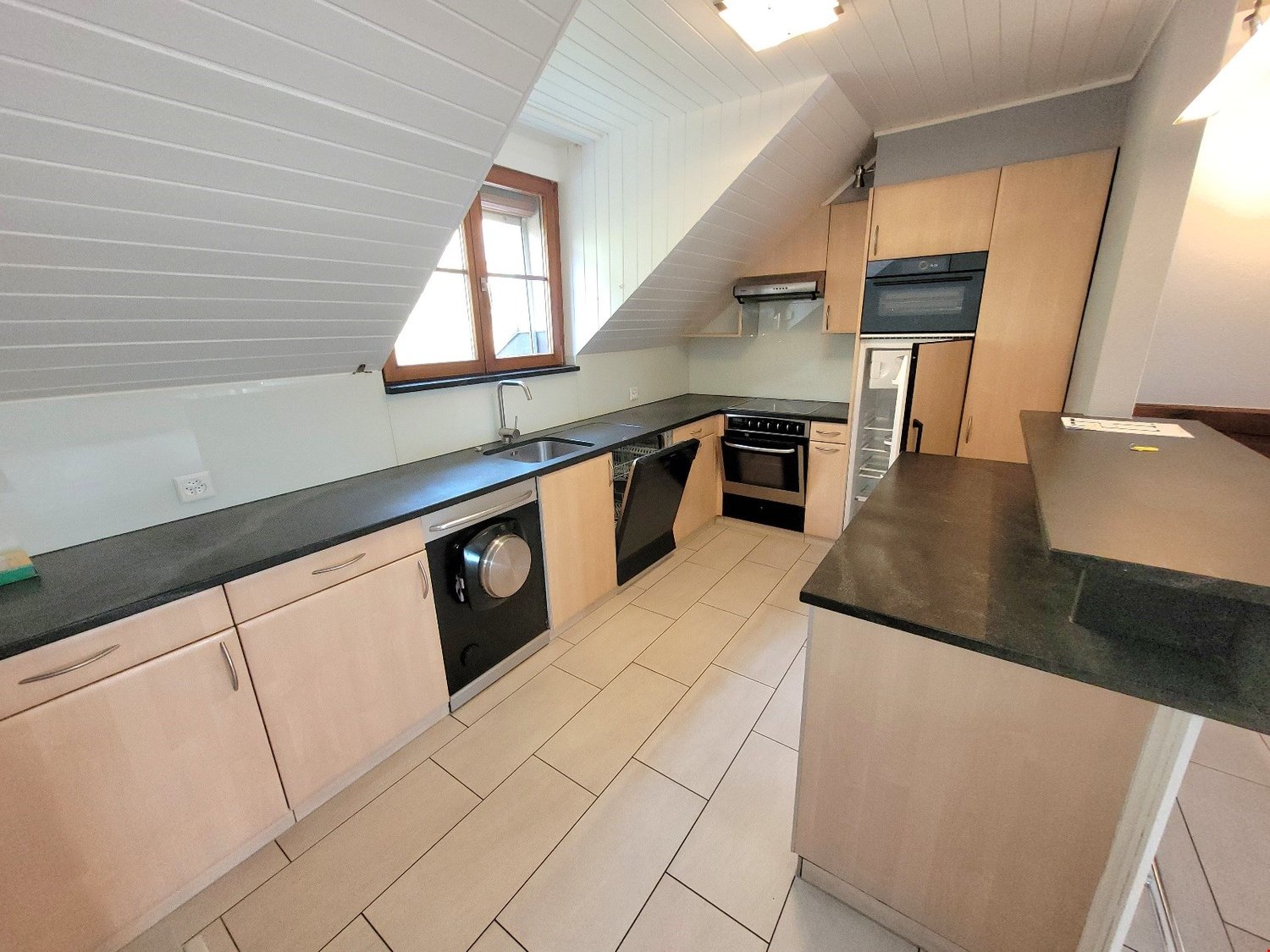 Spacious kitchen with light wood cabinets, black countertops, built-in oven and dishwasher, and a large window providing natural light
