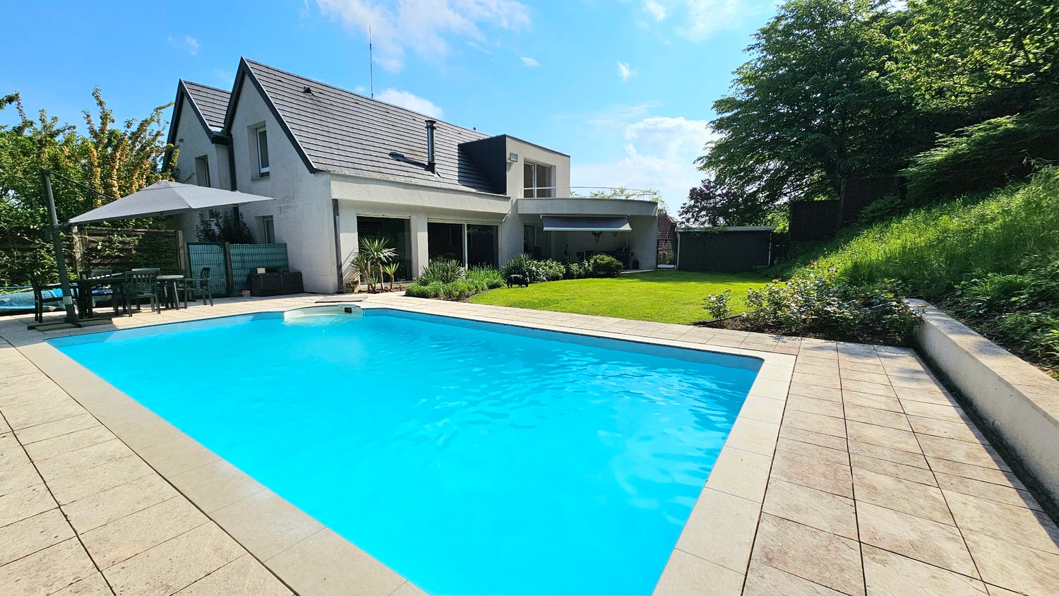 Two-story modern house with a swimming pool, patio, and landscaped garden. The house has a slanted roof and large windows. There is a balcony or terrace visible on the upper floor.
