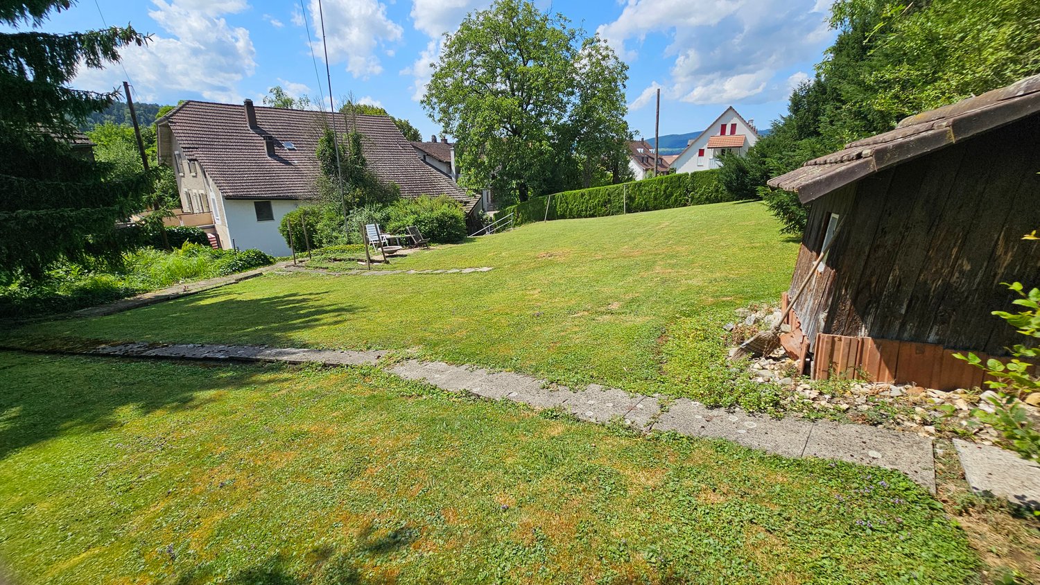 Two-story house with a tiled roof, surrounded by a lush green lawn and trees. There is a wooden shed or structure visible in the backyard.