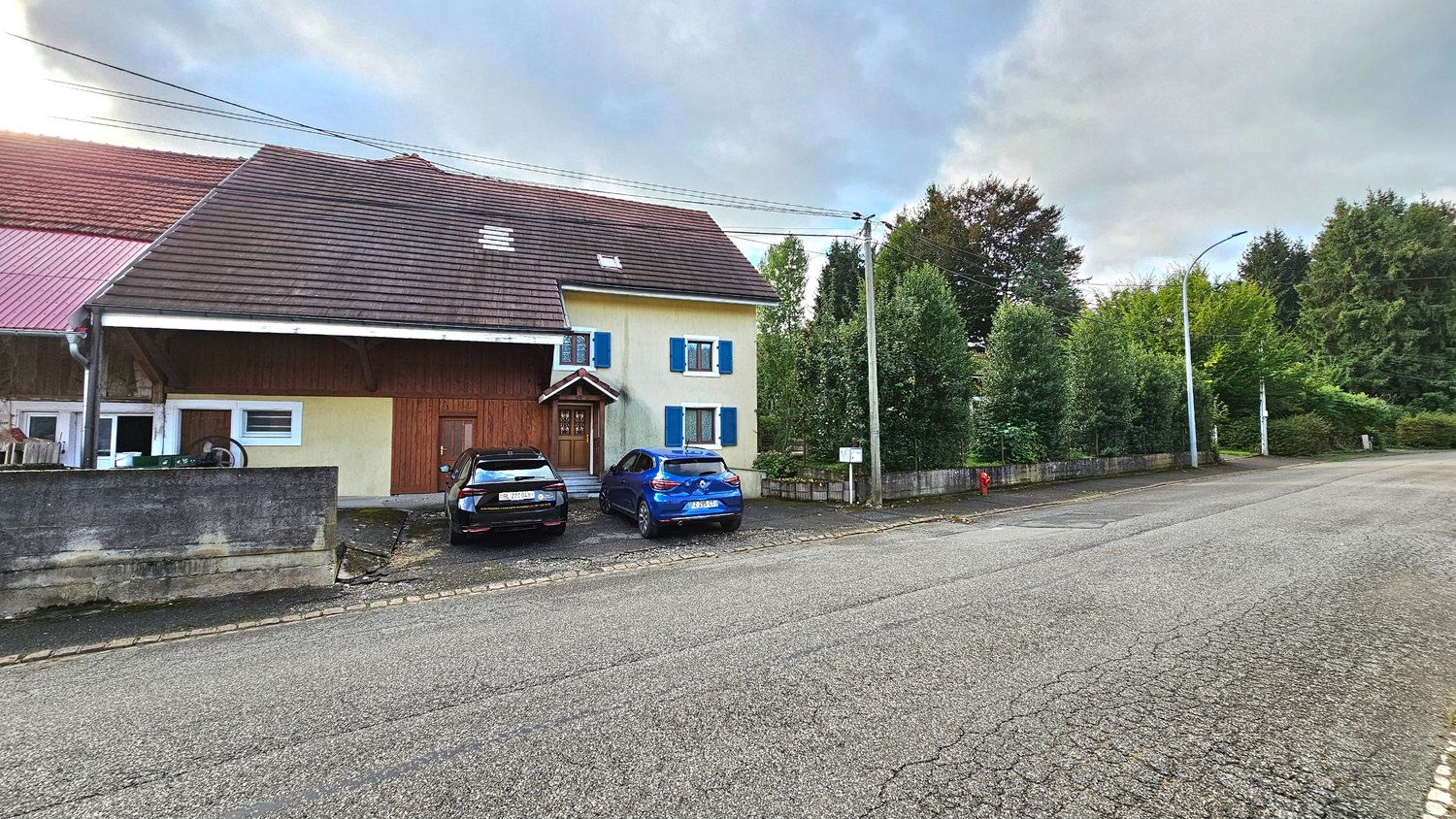 Traditional village house, brown wooden doors, blue shutters, cars parked in front, small garden