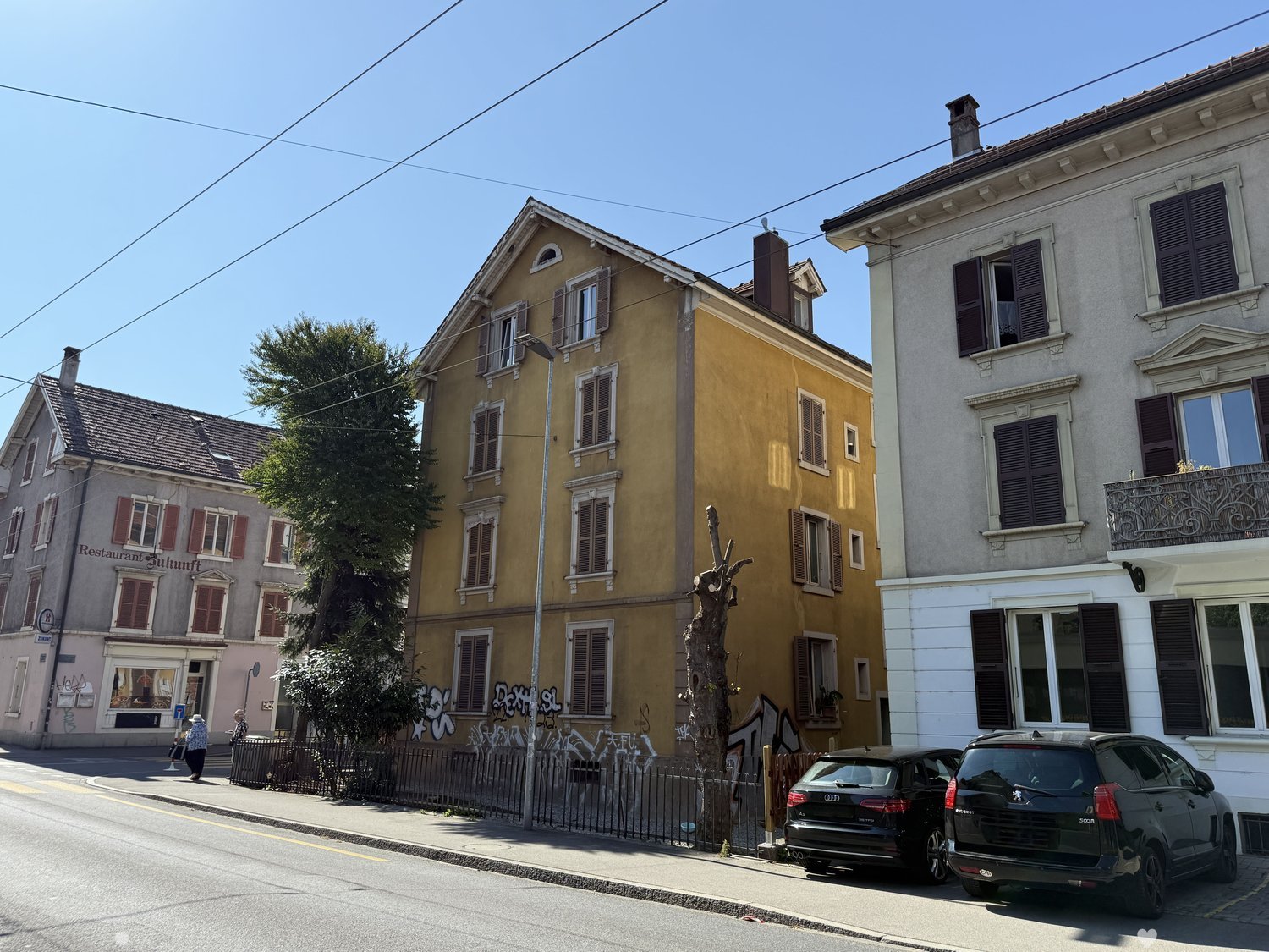 3 story yellow building with balcony and windows, 2 cars parked, 2 other buildings visible