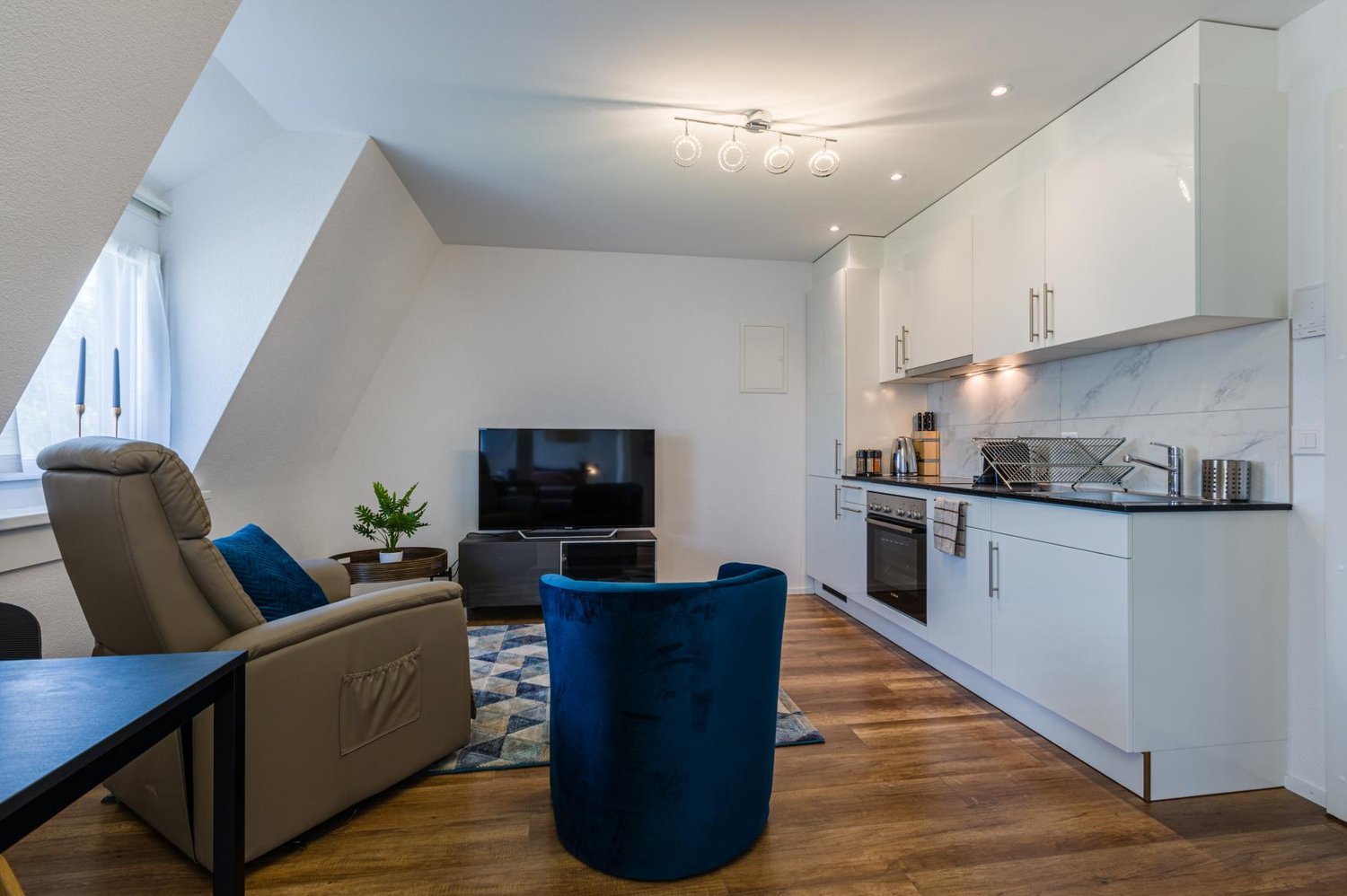 open layout with living room and kitchen area, TV, armchair, rug, modern kitchen, tiled flooring, white cabinets, oven, sink