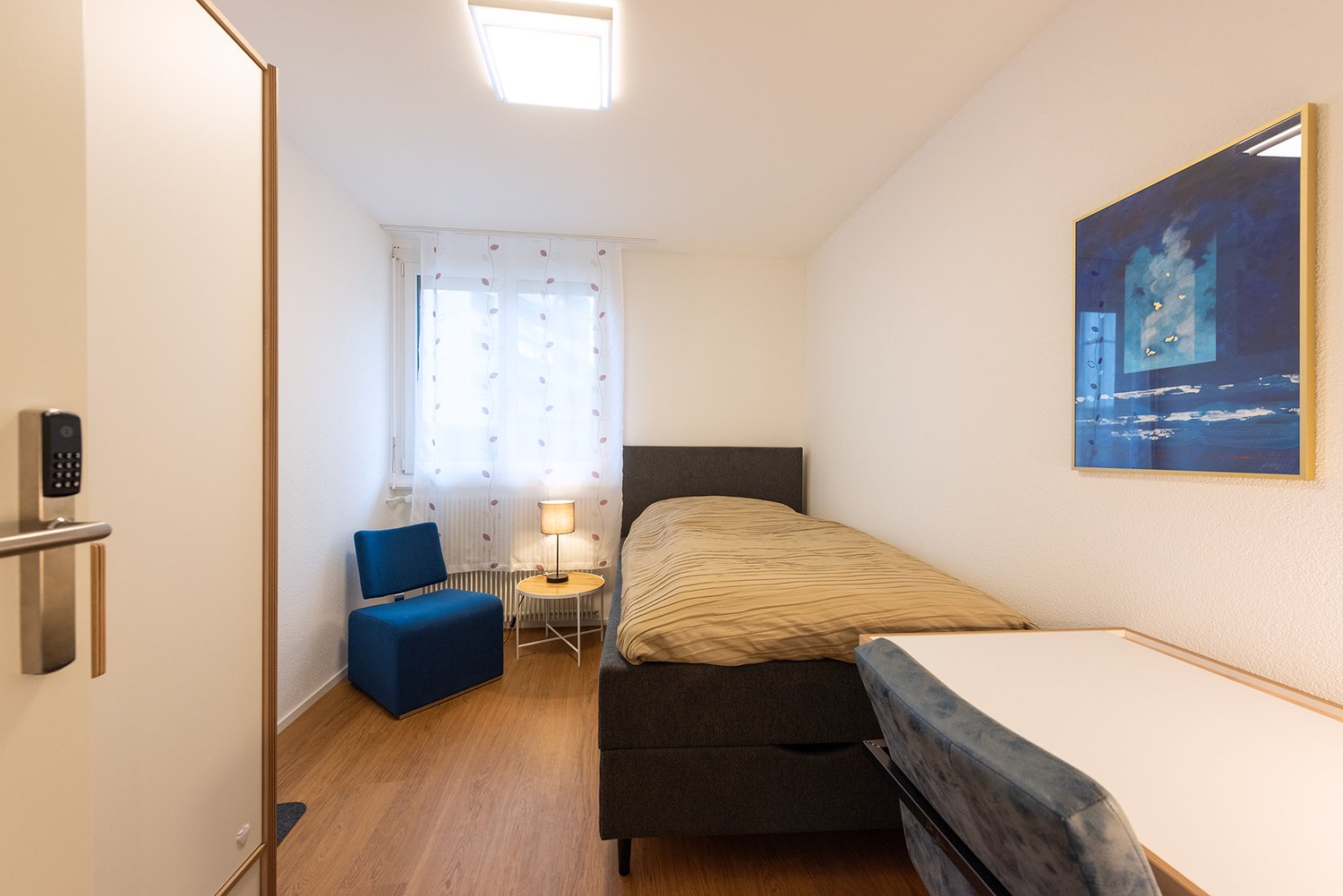 Single bed room with wooden floors, small desk with chair, small blue chair, lamp, white wall, white curtains