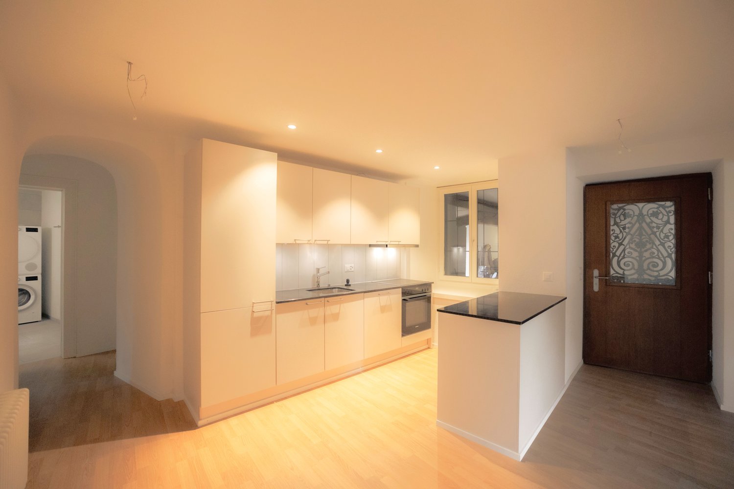 Modern kitchen with white cabinetry, black countertops, wooden floor, and integrated appliances. A large sink with a faucet and oven are visible.