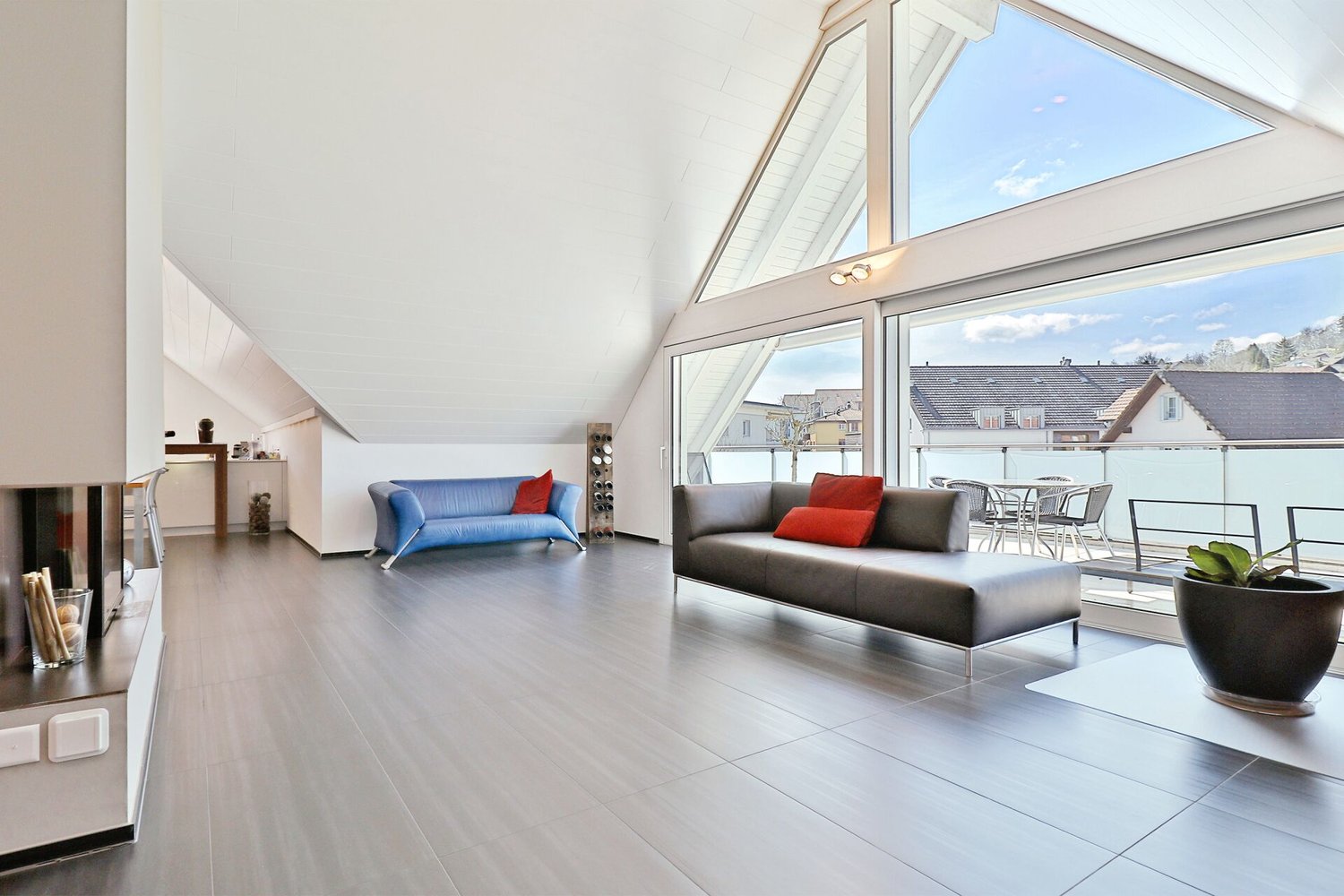 spacious living room with two sofas, fireplace, tiled floor, balcony with table and chairs, sliding doors to balcony, potted plant, large glass windows