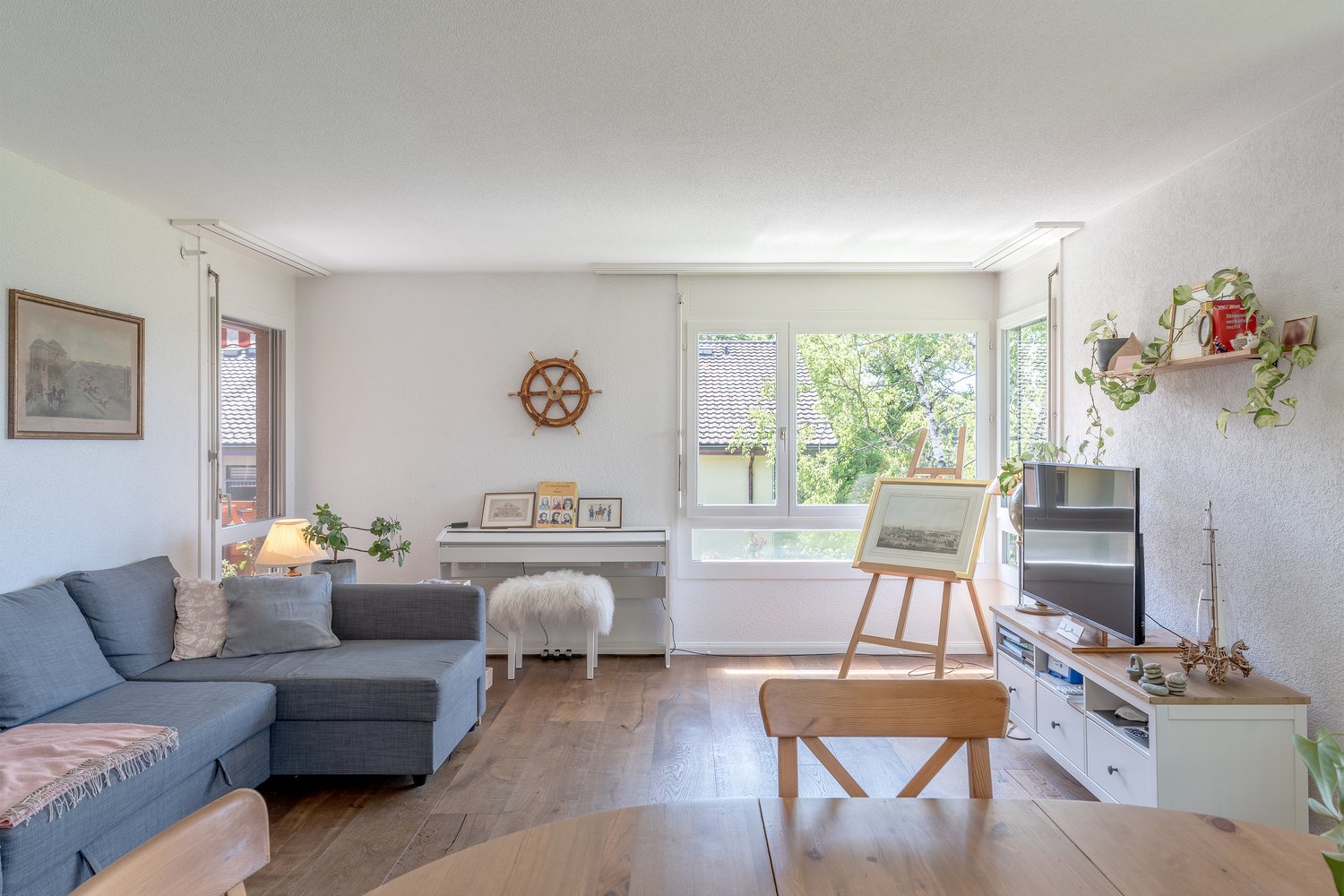 Spacious living room with large windows, grey sofa, wooden furniture, artwork, and potted plants. The room has a cozy and modern feel with a balcony or terrace visible through the windows.