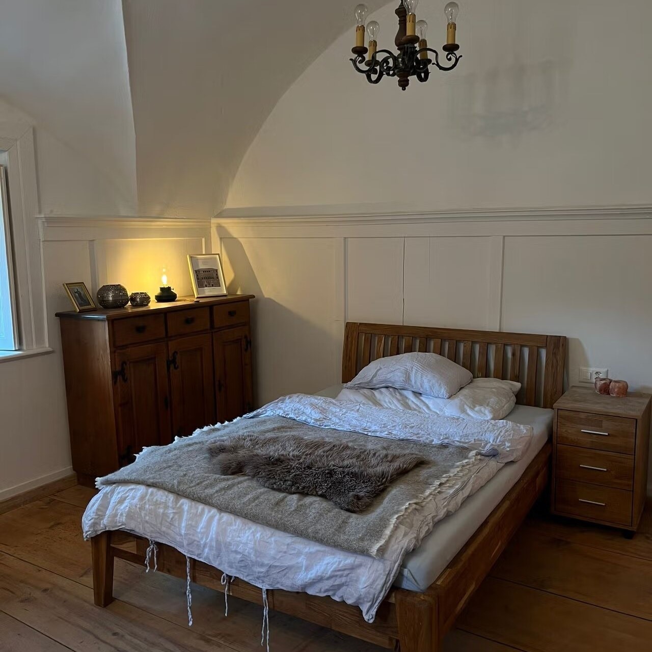 Cozy bedroom with wooden furniture, including a bed frame, dresser, and nightstand. The room has a chandelier overhead and a fireplace visible in the background.