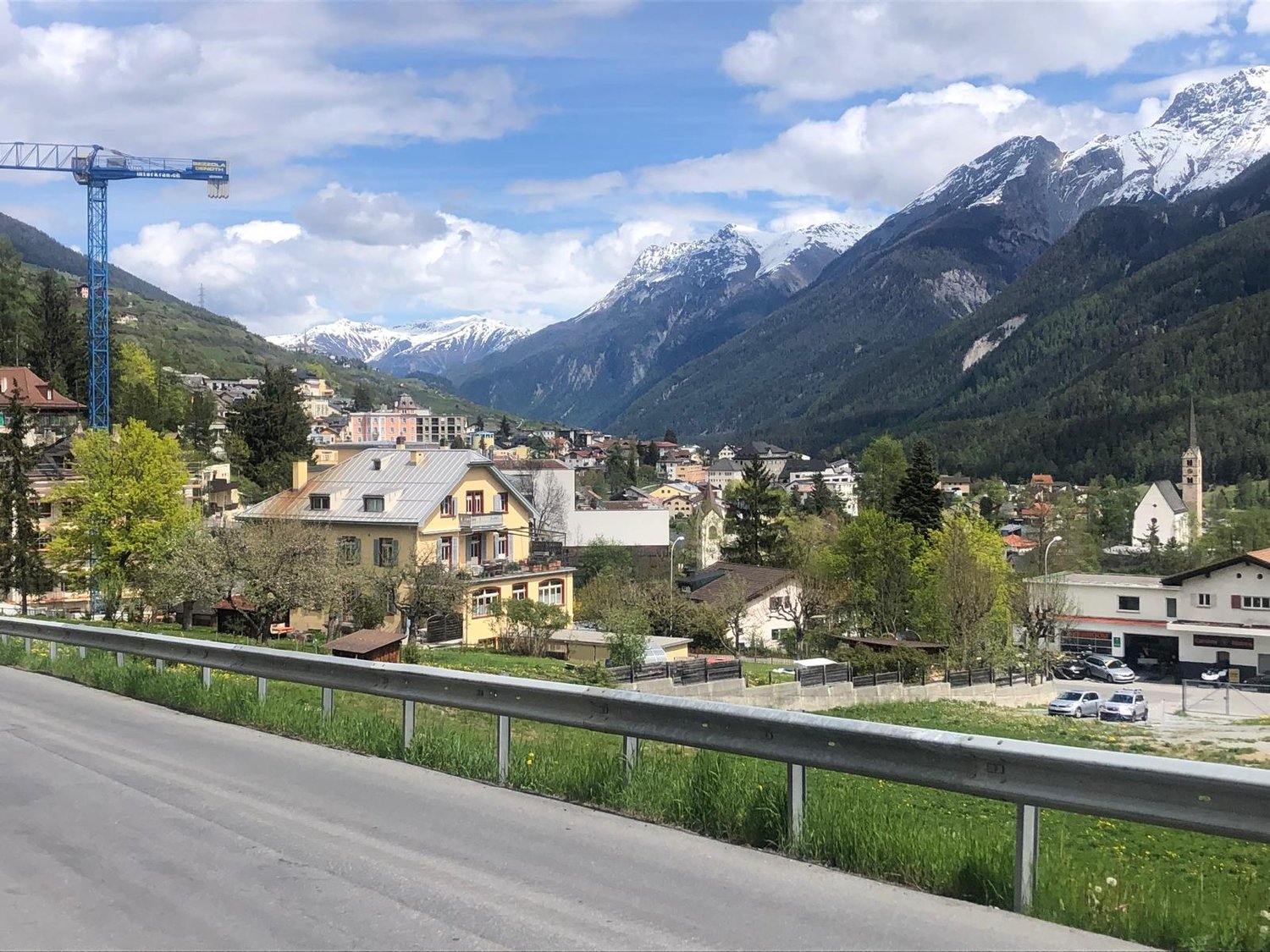 Mountain town with houses, mountains, road, cars, crane, trees, grass, sky