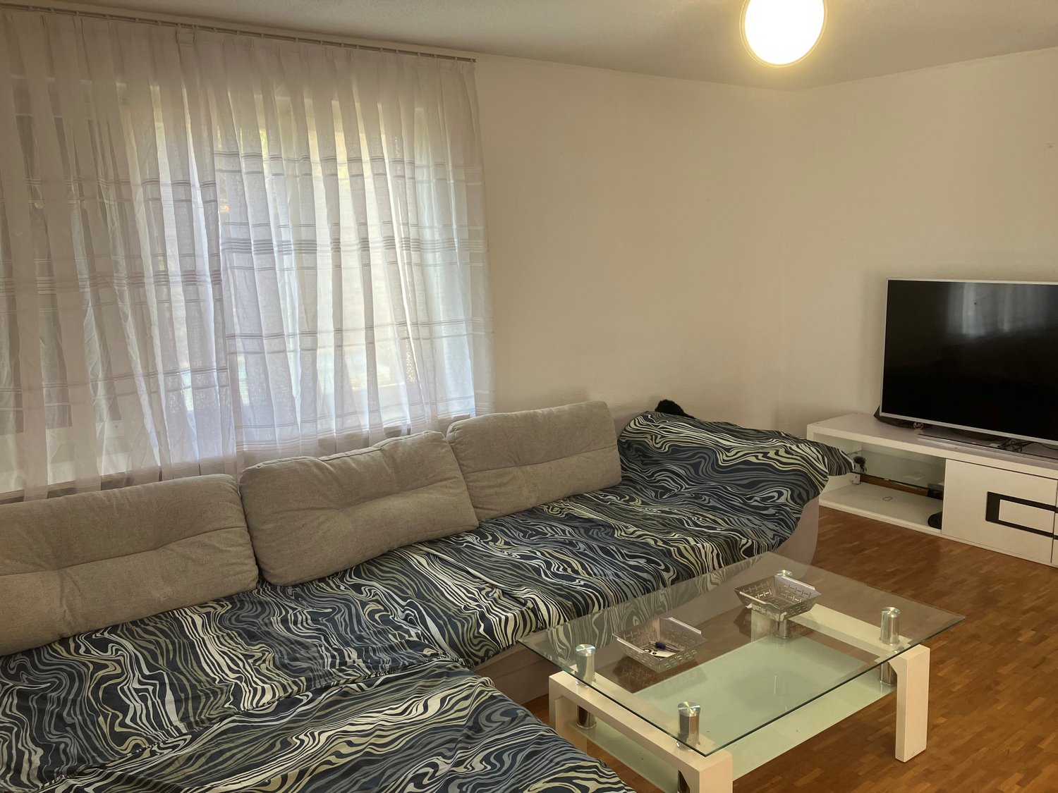 L-shaped sofa, glass coffee table, flat screen TV, white curtains, wooden floor