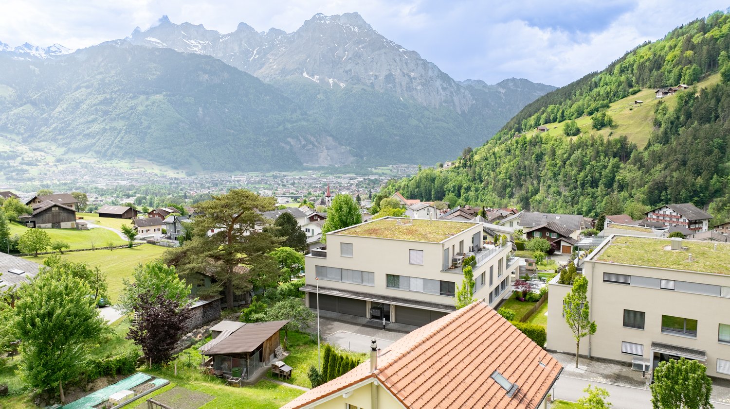 The image shows a picturesque mountain landscape with a small town nestled in the valley below. The town is surrounded by lush green forests and rolling hills, with snow-capped peaks in the distance. The buildings in the town appear to be a mix of traditi
