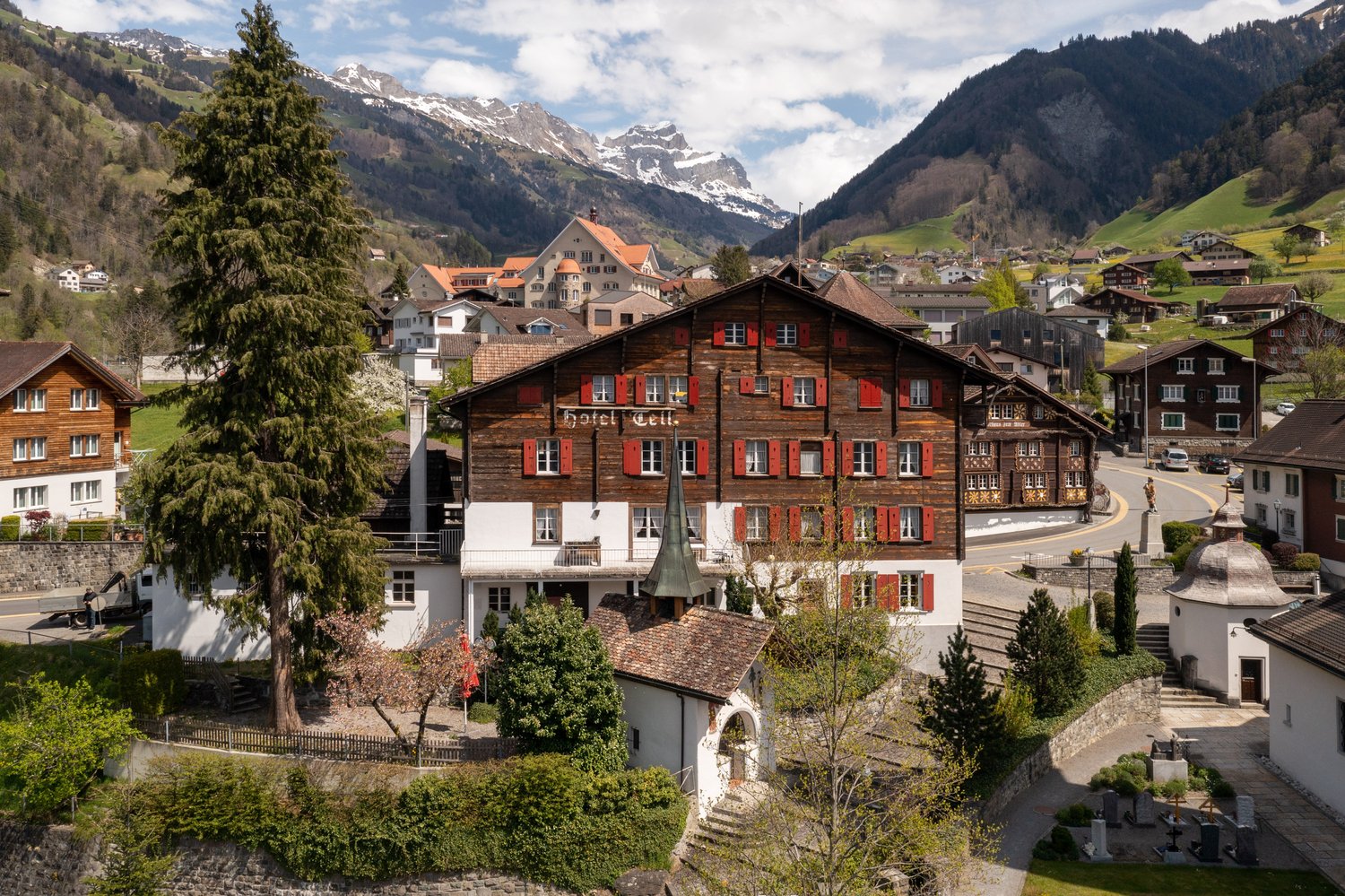 Picturesque alpine town with traditional wooden buildings, a church steeple, and surrounded by snow-capped mountains and lush greenery. The town has a charming, historic feel with a central square and balconies on many of the buildings.