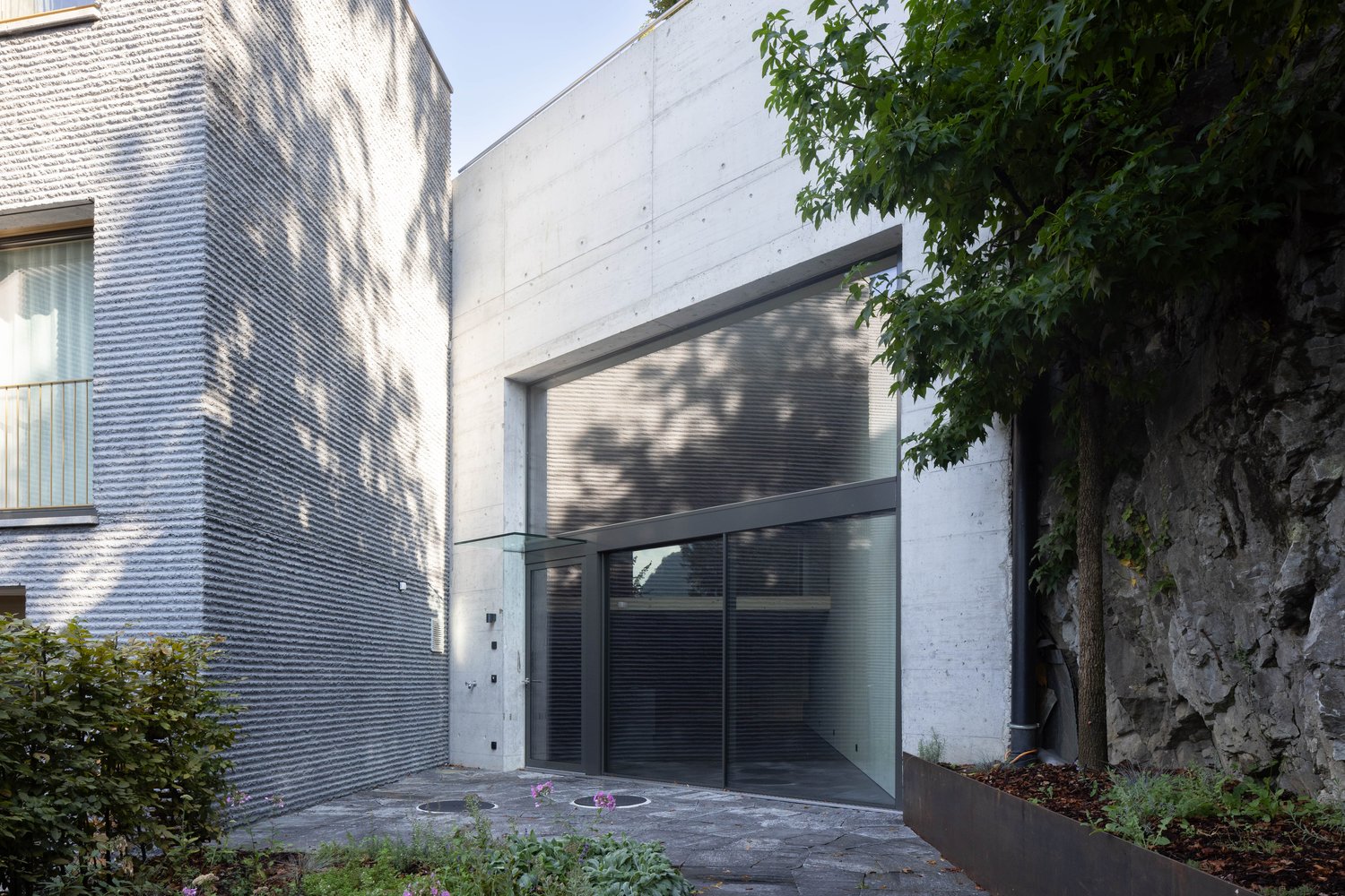 The image shows a modern, two-story building with a concrete exterior and large glass windows. There is a garage door visible on the ground floor, indicating the presence of a garage. The building is surrounded by trees and greenery, giving it a natural,