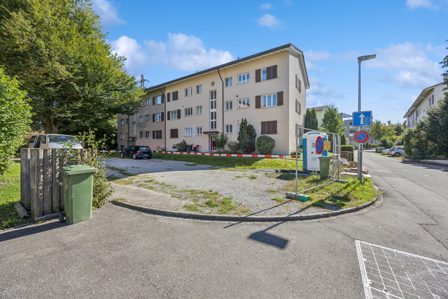 4-story building, gray, fenced entrance, multiple windows, cars parked