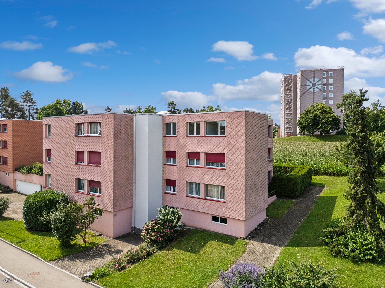 A modern pink residential building with glass windows, set in a suburban landscape with grass and trees. Several plants and shrubs are arranged in the front. In the background, there is a larger building with a tower.