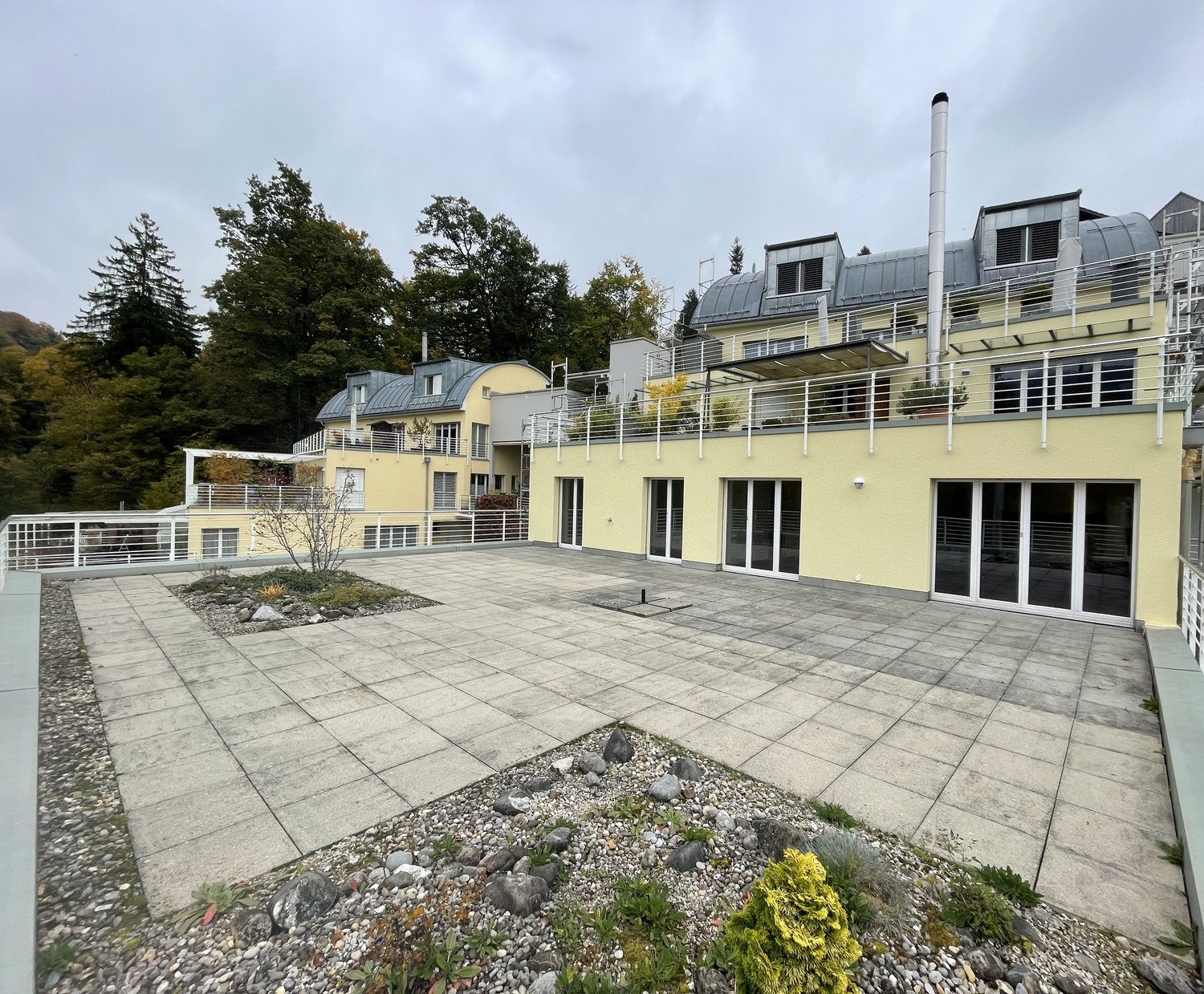 Terrace with paved floor, metal handrails, surrounding trees, yellow building with many glass doors, balcony with metal railing