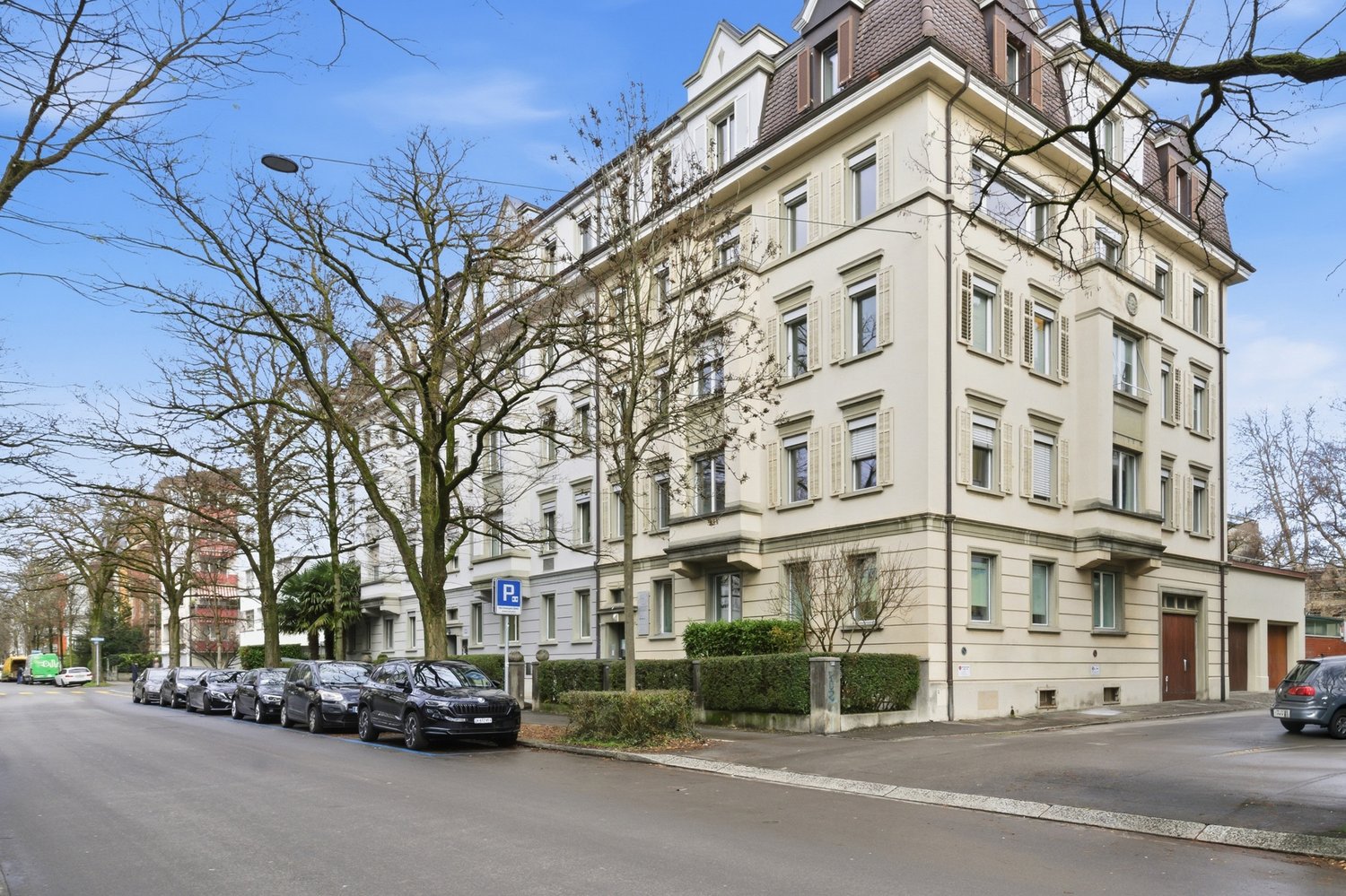 A large white building with multiple windows and balconies, surrounded by trees and bushes, with cars parked in front.
