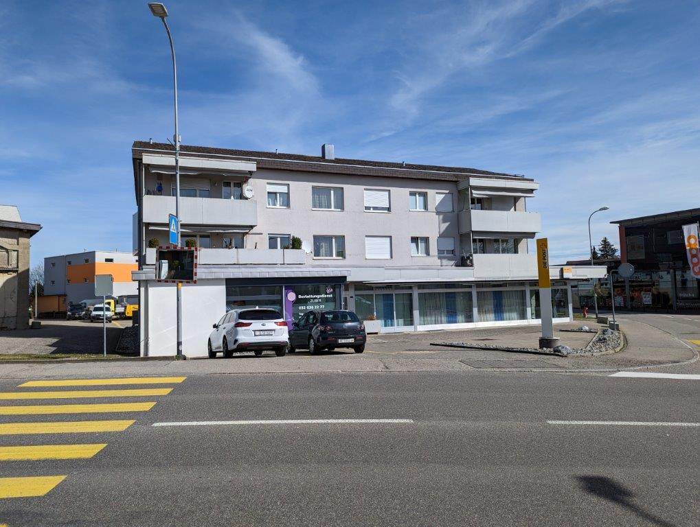 A multi-story apartment building with a commercial space on the ground floor. The building has a modern, rectangular design with white walls and large windows. There are cars parked in front of the building, and a crosswalk is visible on the street.
