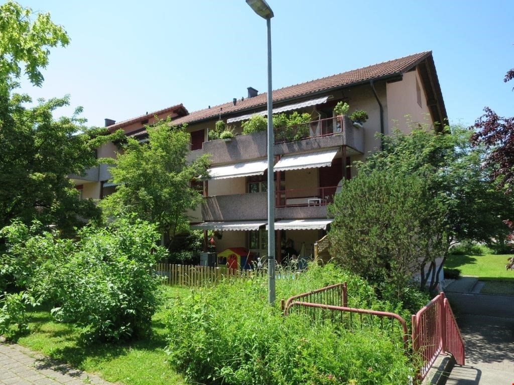 Three story apartment building, red tiled roof, balcony with potted plants, white awnings, red fence, paved walkway, streetlight, fenced yard with plants and trees, playground structure nearby
