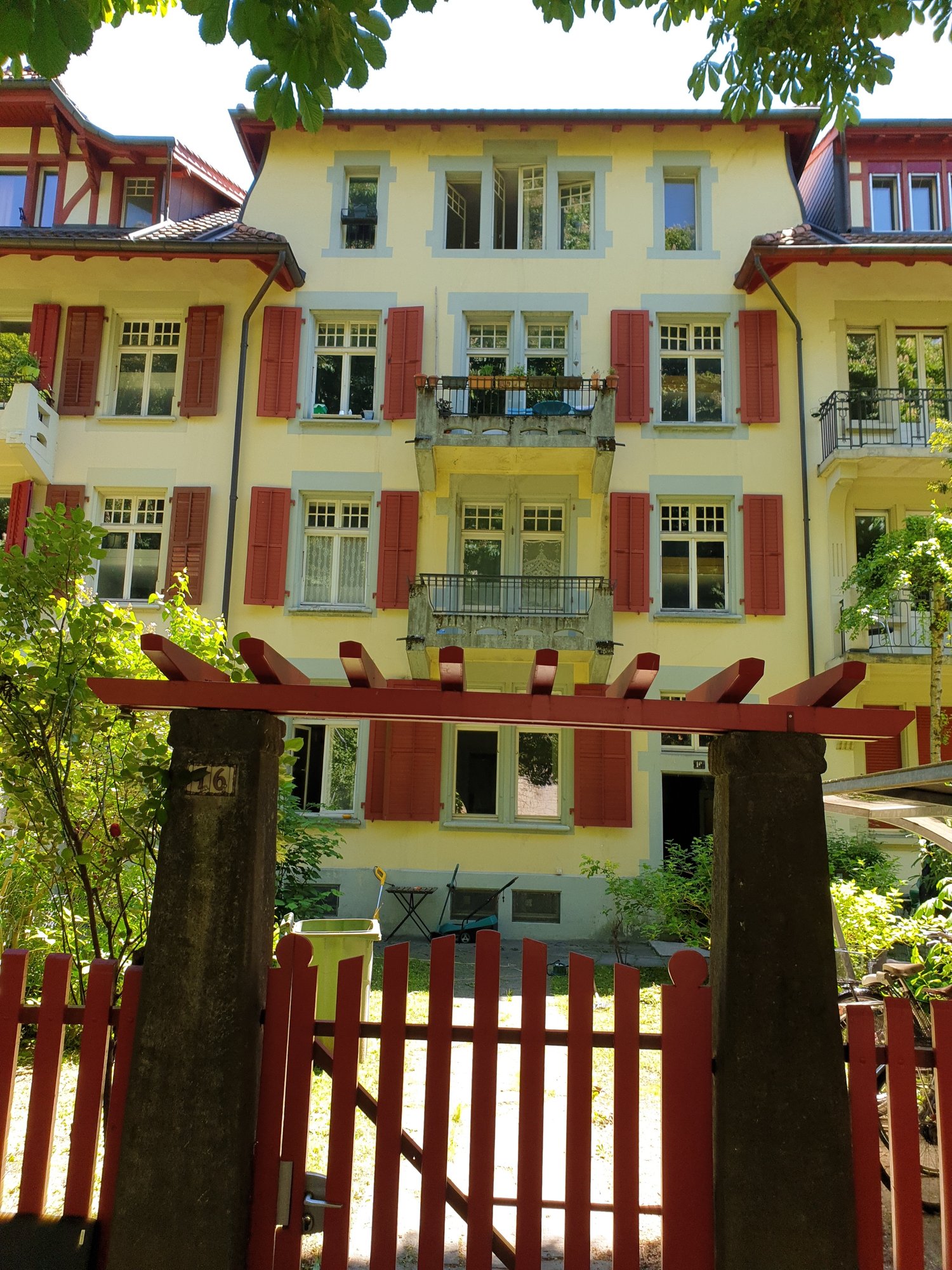 Yellow 3 story house, red shutters, balconies on upper floors, green trash can, red gate, plants and trees