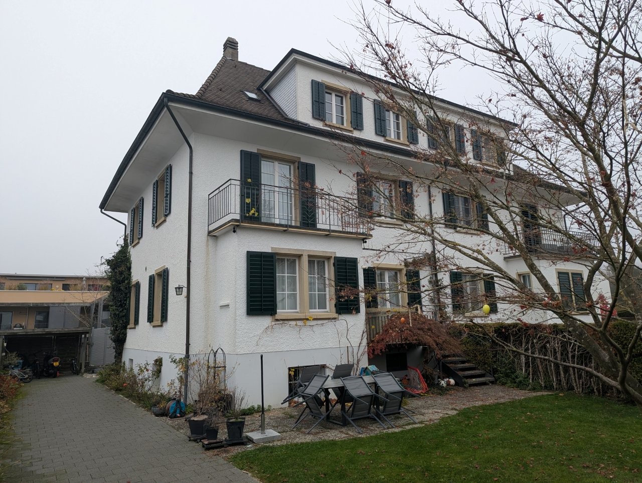 3-story building, white painted, black gutters, black metal balcony, green window shutters, brick driveway, garden, small plants