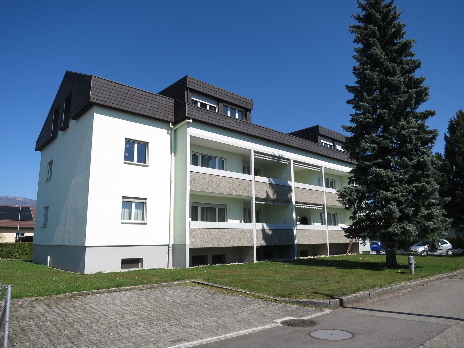 white and gray exterior, multiple floors, balconies with awnings, glass windows, ground floor and upper floor windows, black roof, paved sidewalk, car parked in front of the house