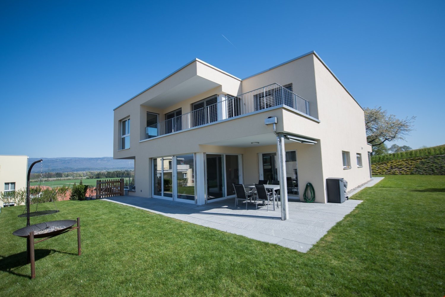 Two-story modern house with a balcony, large windows, and a patio area with outdoor furniture. The house has a well-manicured lawn and is situated in a scenic location with a view of the surrounding landscape.