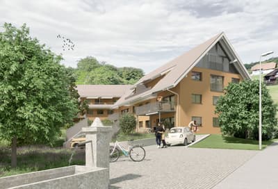 3-story apartment building with a slanted roof, surrounded by trees and greenery, with a paved courtyard and parking spaces in front
