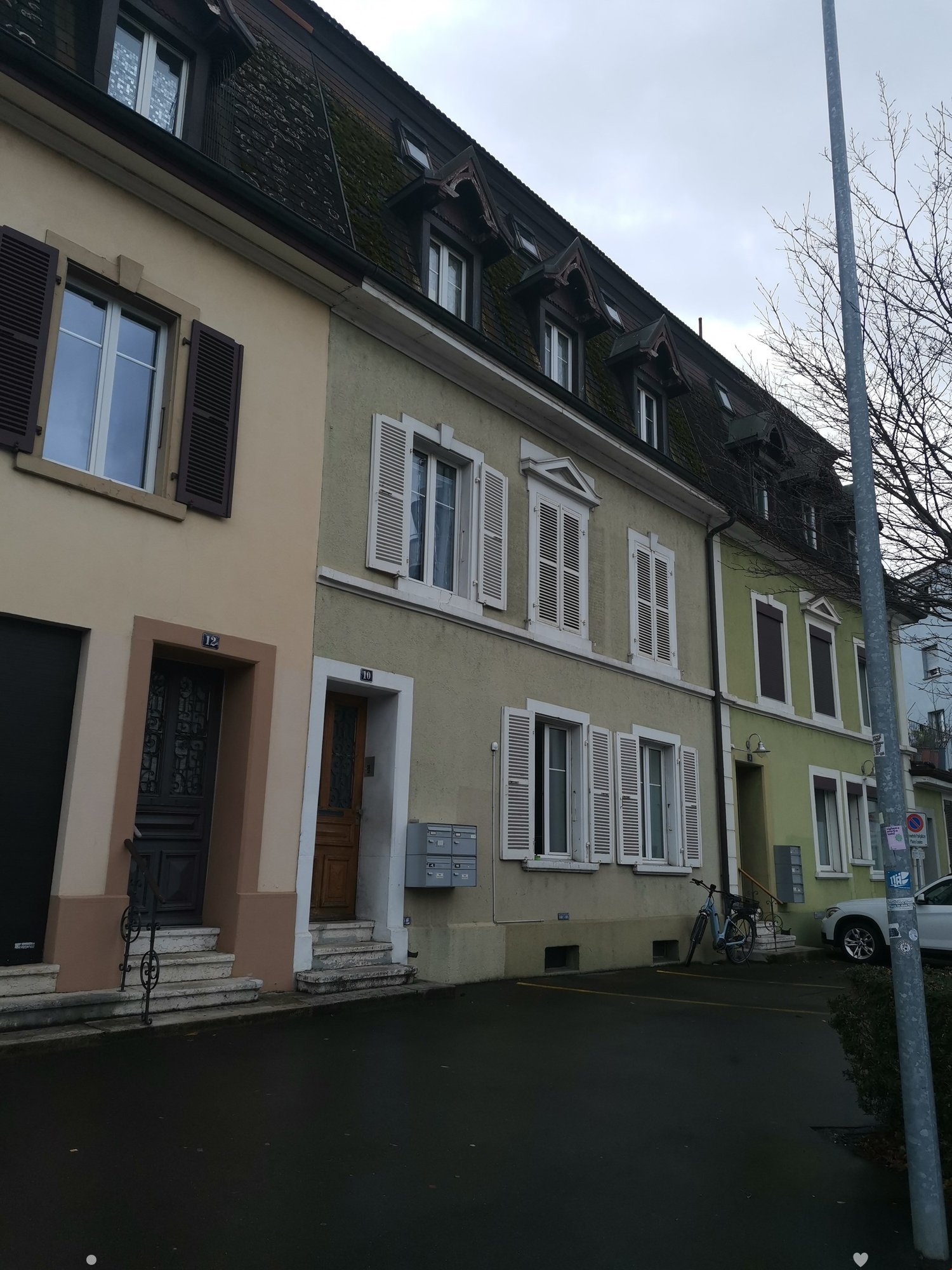 Multi-story apartment building with a green exterior, white shuttered windows, and a slanted roof with dormers. There is a bicycle parked in front of the building.