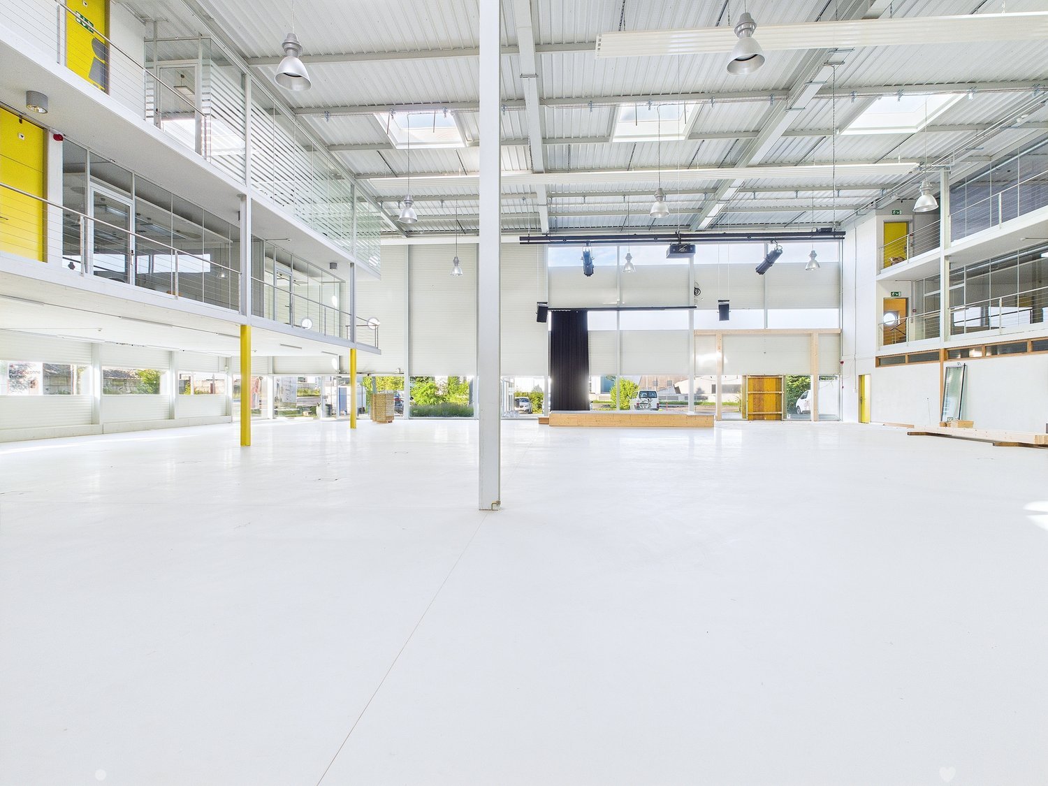 Large open commercial space with high ceilings, white walls, and yellow accents. The space has a stage-like area and appears to be a multipurpose event or exhibition hall.