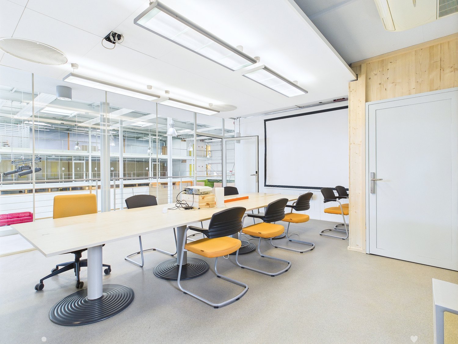 Modern office space with large conference table, yellow and black chairs, projector screen, and glass walls providing natural light