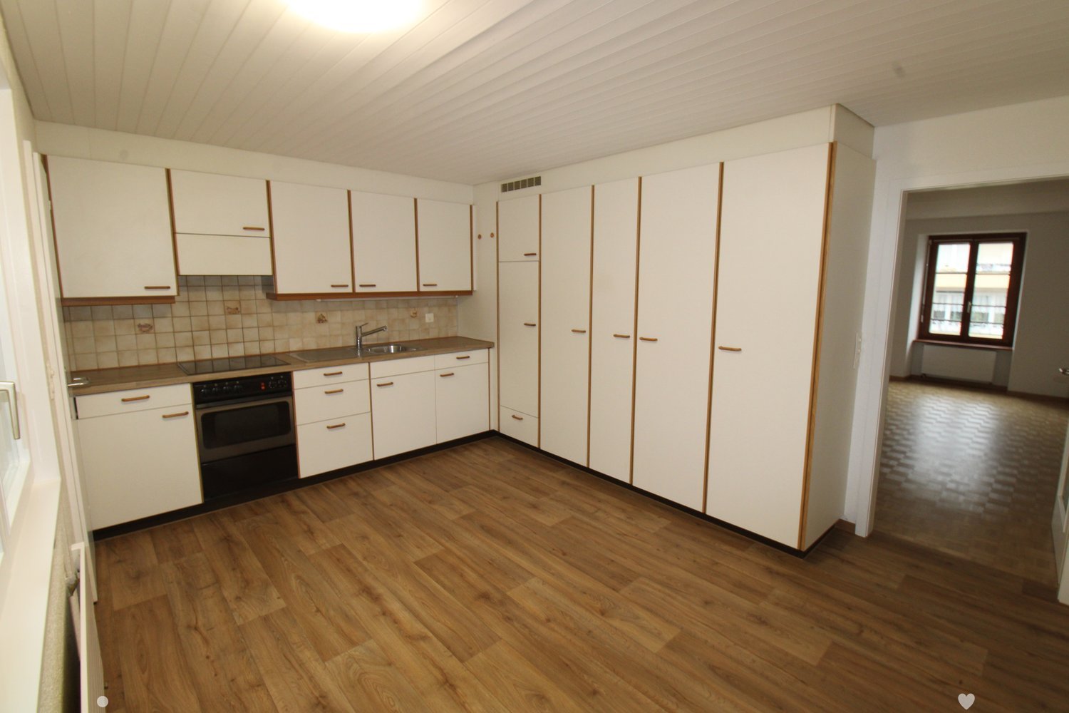Empty kitchen with wooden floor, white cabinets, black appliances, sink, window.