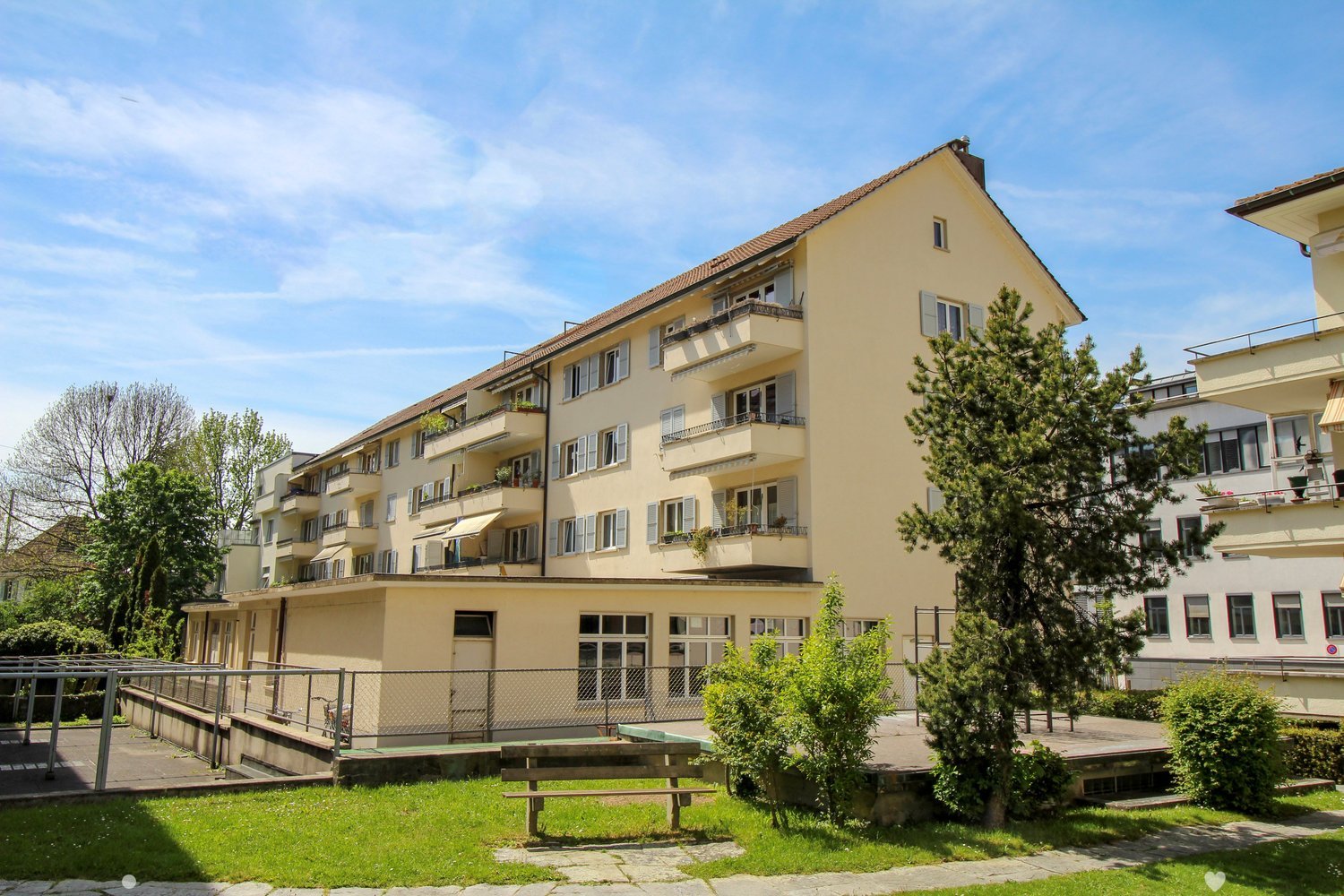 Apartment building with many balconies, multiple floors, fence surrounding the area, trees, and green space
