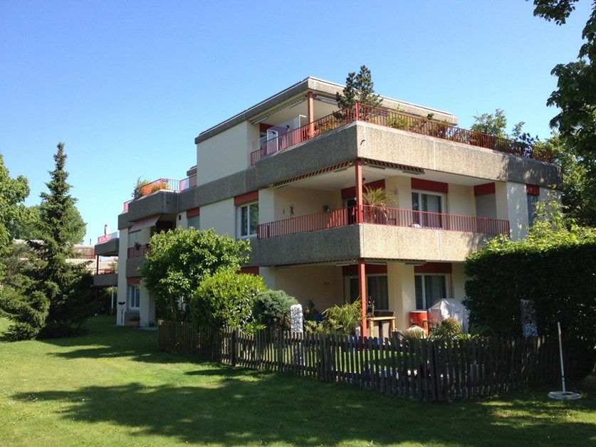 Two story house, white and red walls, large windows, balcony on each floor, surrounded by greenery
