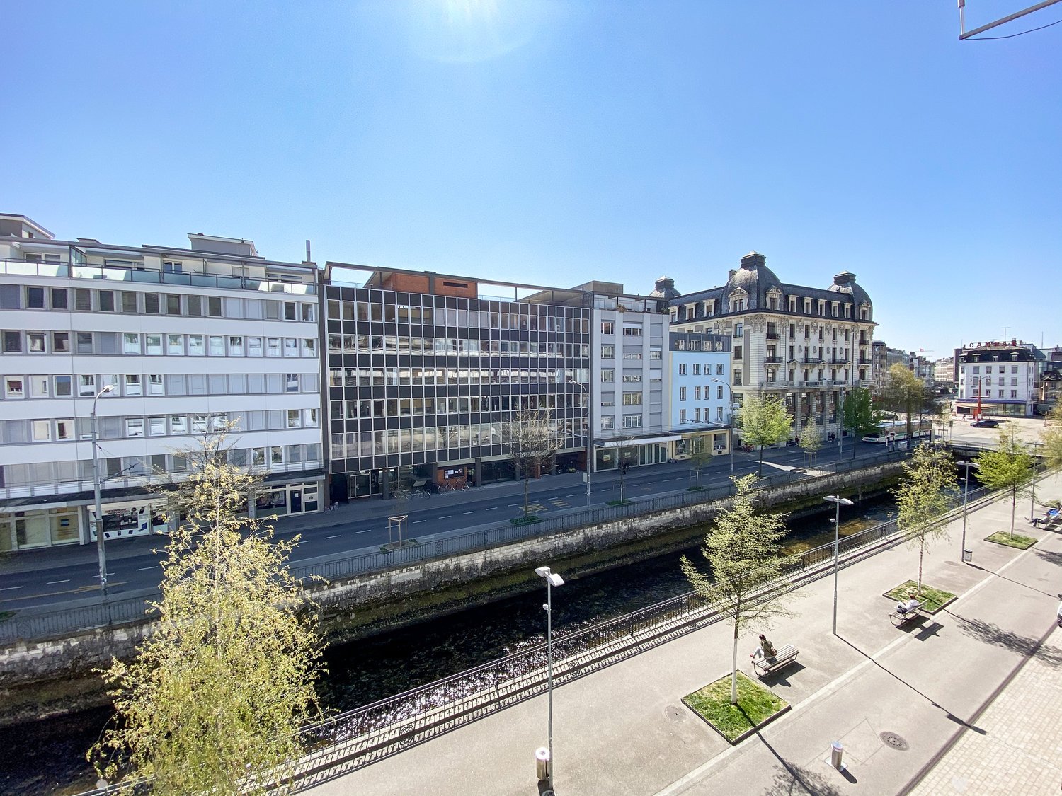 Aerial view of a city with multiple buildings, a river, and a road with streetlights and benches.