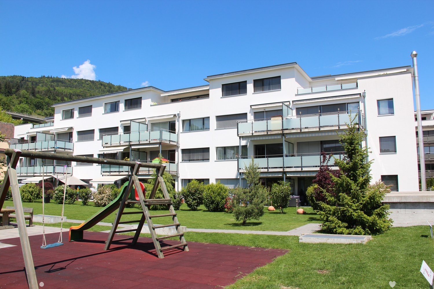 Multi-story apartment building with white exterior, balconies, and a playground area with swings in the foreground. The building is surrounded by green landscaping and trees.