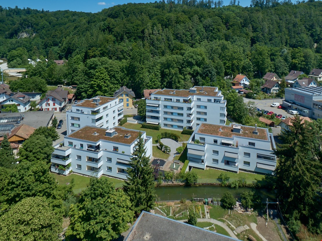 Group of apartments on a hilly location, surrounded by trees and grass, with a river in the foreground