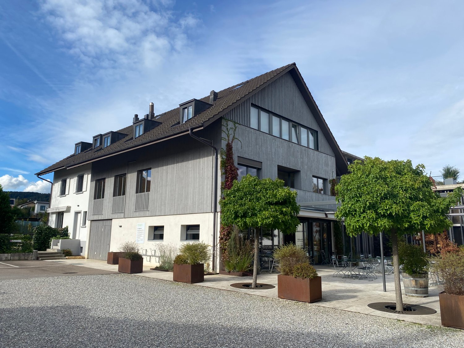 3-story building with a gray exterior, large windows, and a balcony. There are trees and planters in the front of the building, and a paved outdoor seating area.