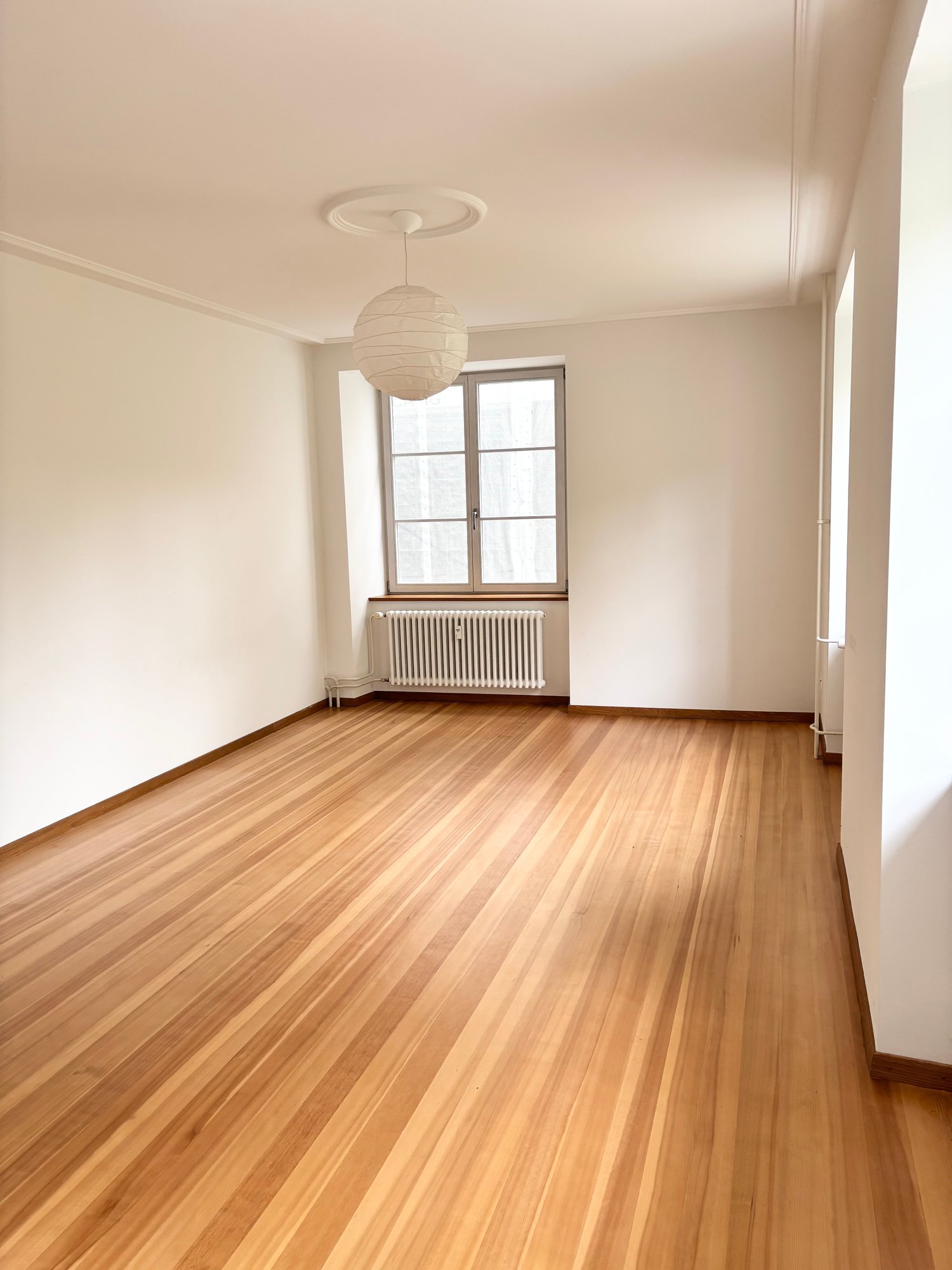 Empty room with wooden floor, white walls, and radiator. Window with glass and radiator.