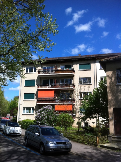 4 stories, balconies, beige walls, grey windows, red awnings, hedges, small garden, multiple cars parked in front
