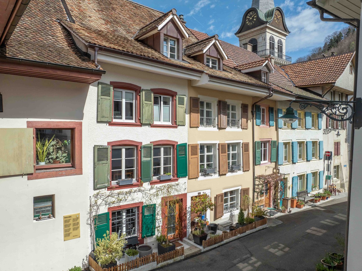 This image shows a charming, historic town square with colorful, traditional Swiss-style buildings. The buildings have red tile roofs, white walls, and decorative shutters in various colors like green, red, and blue. There is a clock tower in the backgrou
