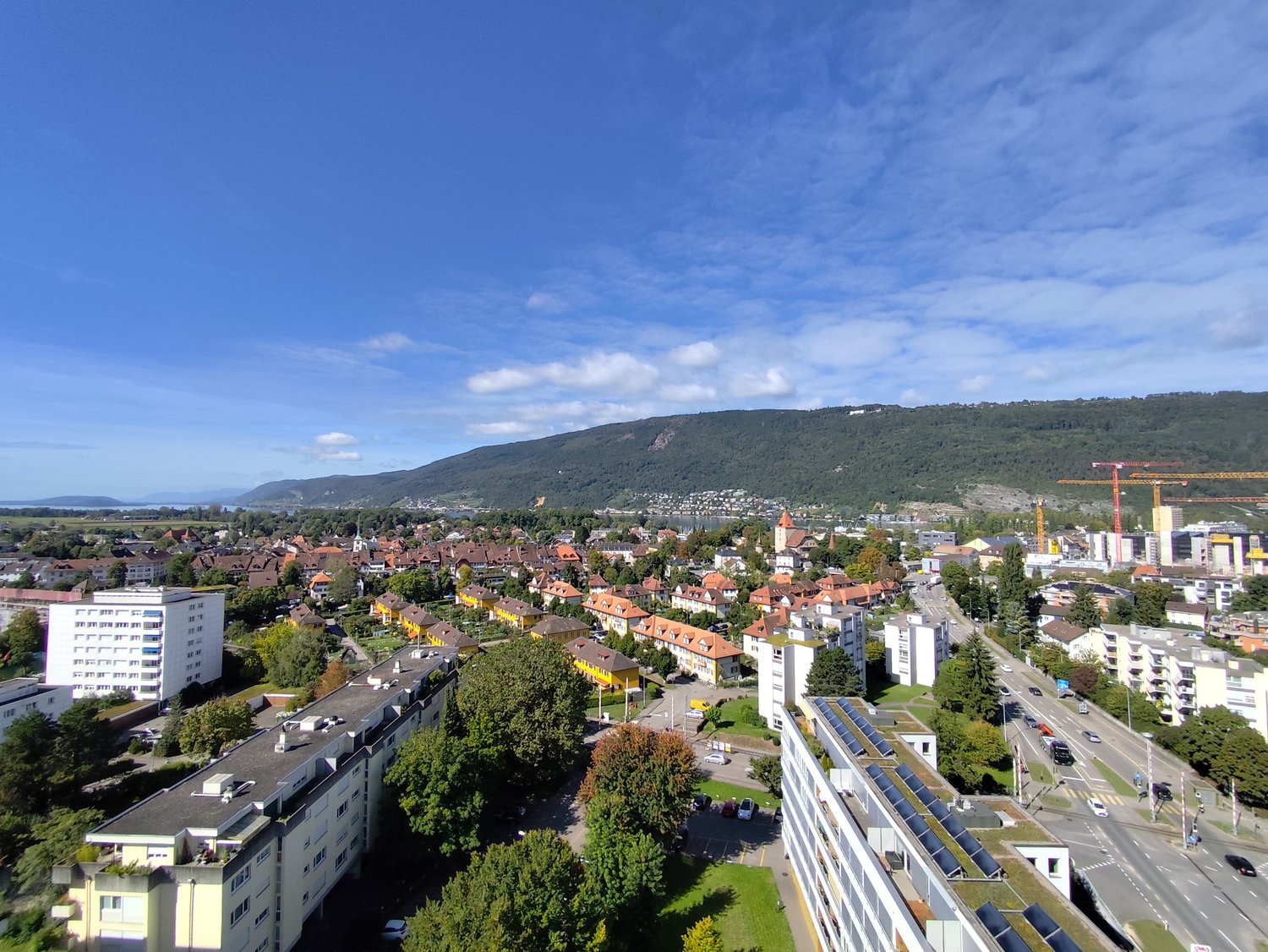 Aerial view of an urban residential area with numerous buildings, solar panels, trees, and a road with moving vehicles in the background, with a beautiful view of the sky and mountains