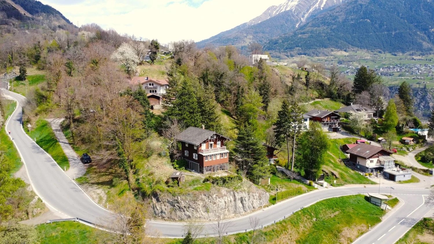 Aerial view of a mountainous, forested area with a small village nestled among the trees. The landscape features winding roads, scattered houses and chalets, and a backdrop of snow-capped mountains.