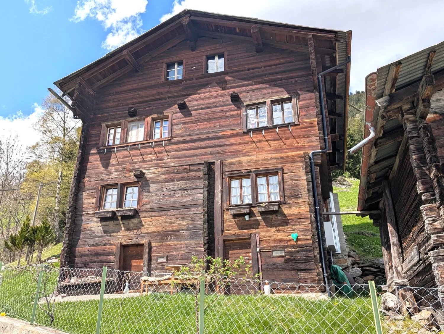 Two-story wooden chalet-style house with a slanted roof, wooden exterior, and multiple windows. The house appears to be located in a rural or mountainous area with a grassy field in the foreground.