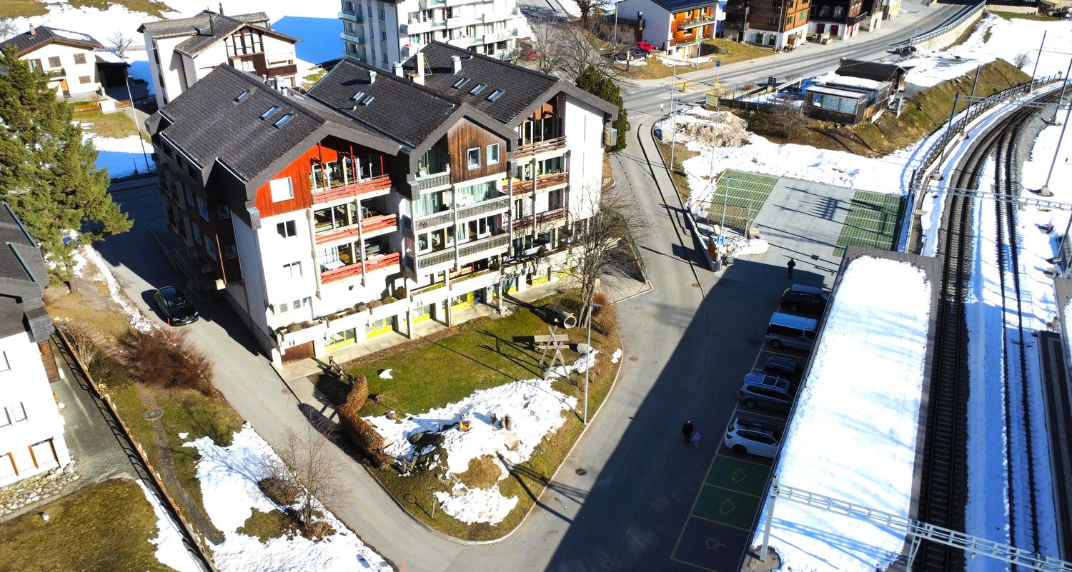 Multi-story apartment building with red and white exterior, balconies, surrounded by snow-covered landscape, with a road and ski lift visible in the background.