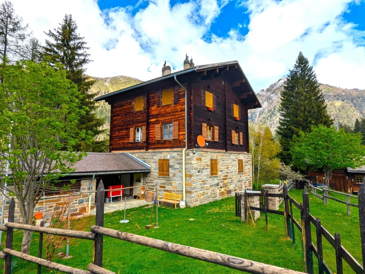 Two-story wooden chalet-style house with stone foundation, surrounded by lush green grass, trees, and mountains in the background. The house has a balcony or terrace on the upper level.