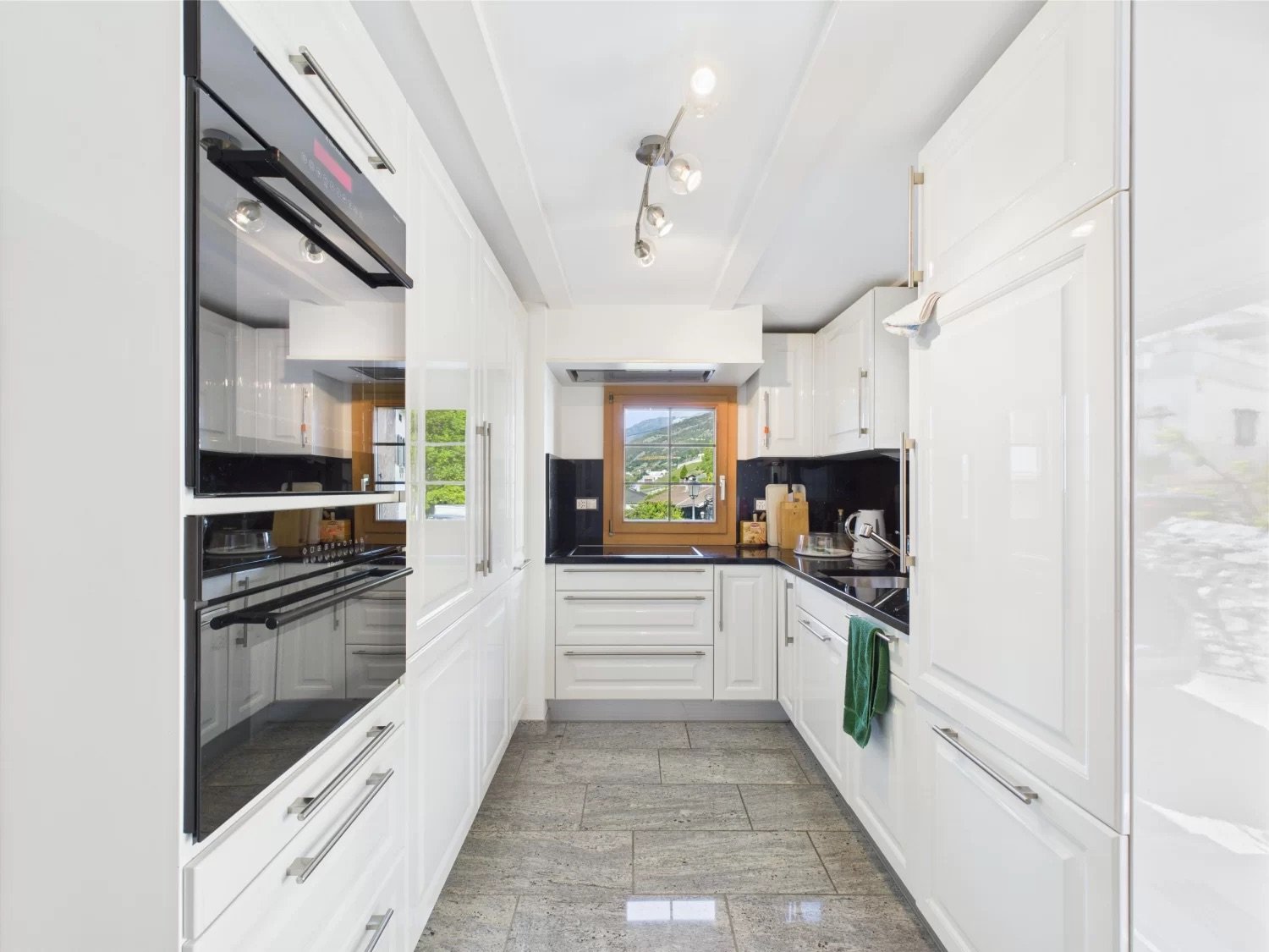 Spacious modern kitchen with white cabinets, black countertops, stainless steel appliances including a dishwasher, and large windows providing a scenic view.